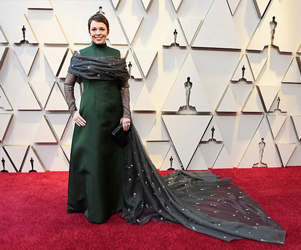 A person in a green gown with a starry shawl on a red carpet at an awards event, geometric backdrop visible.