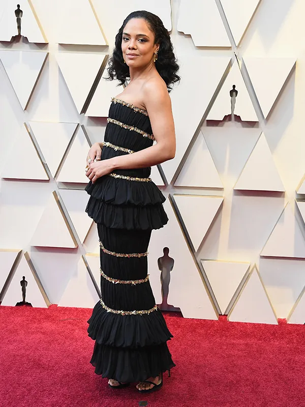 Woman in a tiered black gown with gold accents poses on red carpet at an event.