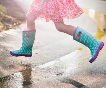 Child wearing colorful rain boots jumps over a puddle on a sunny day.