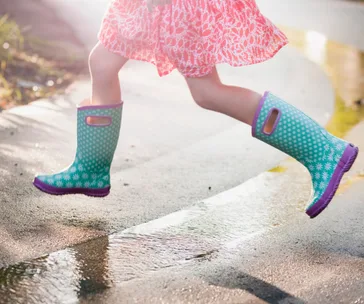 Child wearing colorful rain boots jumps over a puddle on a sunny day.