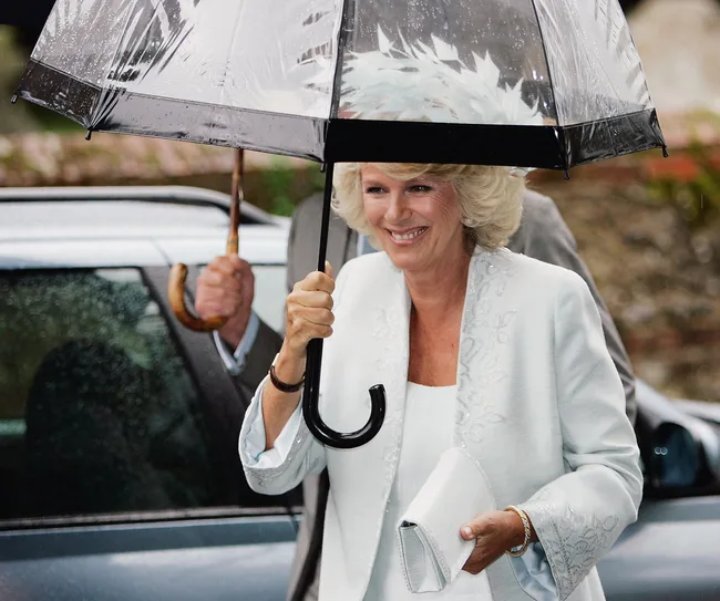 A woman in a light outfit holds a transparent umbrella, smiling outside near a car in the rain.
