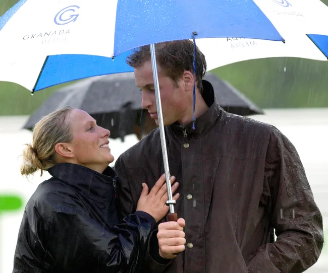 A man and woman smiling under a blue and white umbrella in the rain, wearing dark rain jackets.
