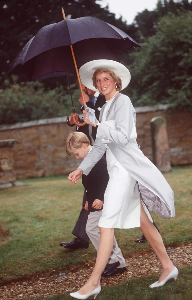 A woman in a white outfit and hat smiles, holding an umbrella, walking with a boy on a grassy path.