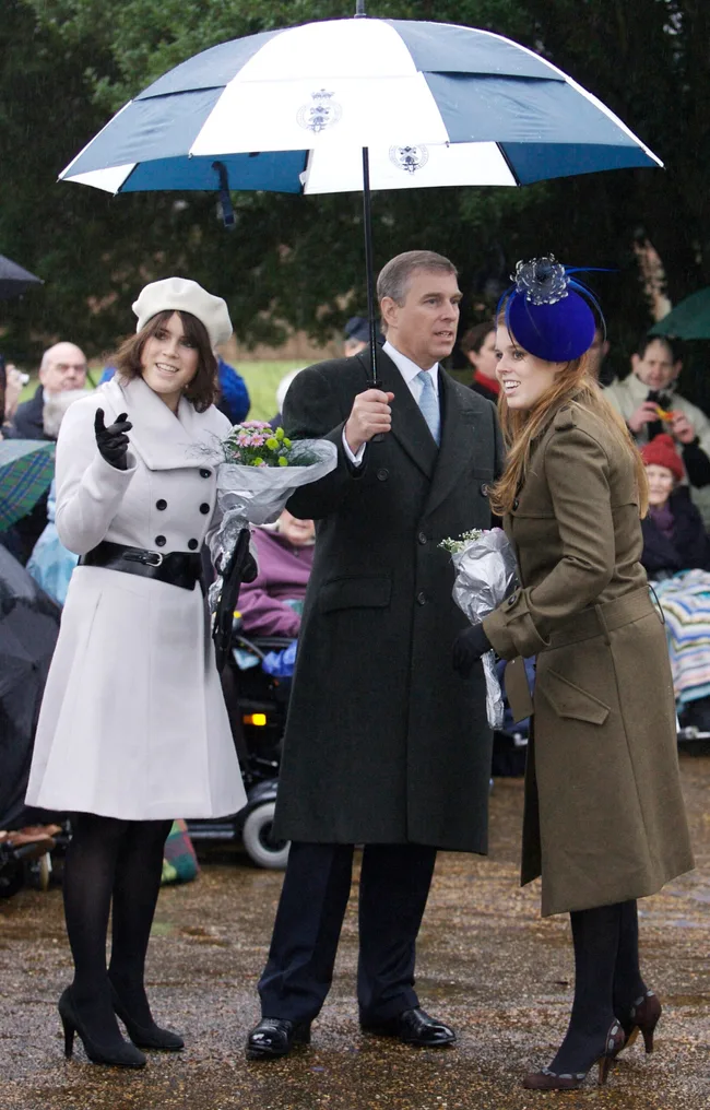 Three people in formal attire stand under an umbrella, holding flowers, while talking in an outdoor setting.
