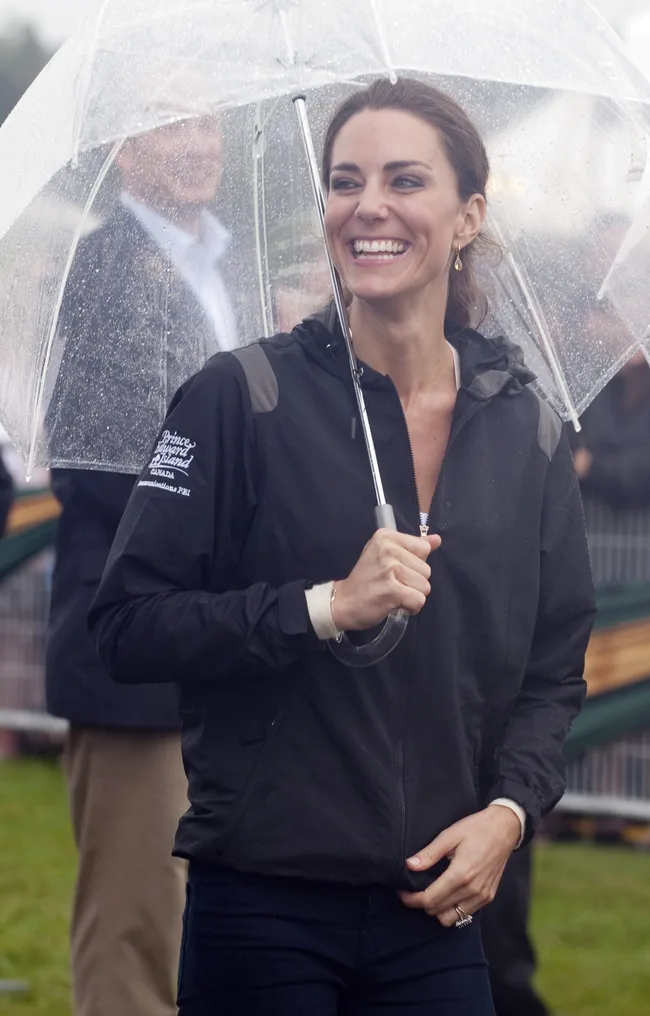 A woman smiling under a transparent umbrella, wearing a black jacket, outdoors in rainy weather.