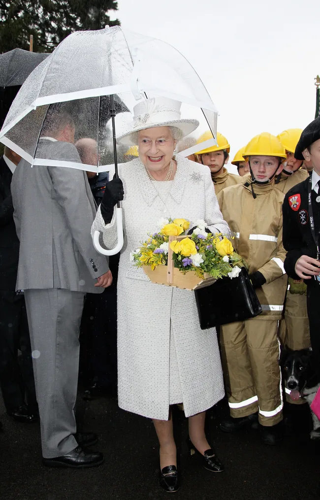 A woman in a white outfit holds yellow flowers under a clear umbrella, surrounded by people in yellow helmets.