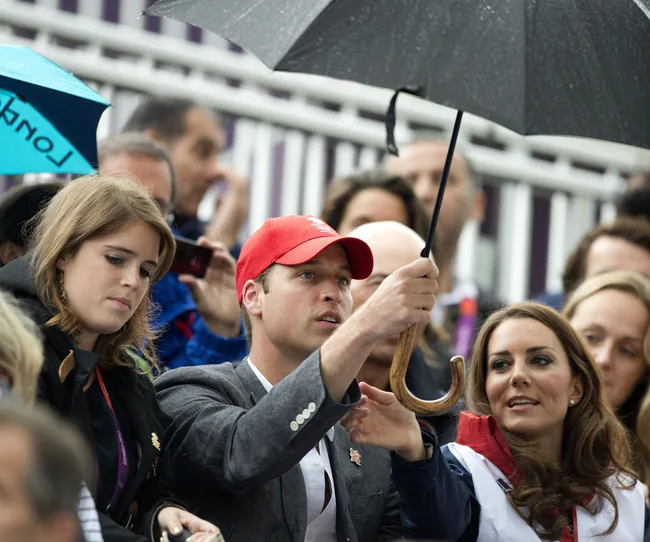 Three people are seated in a crowd, one holding an umbrella. The image is taken at an outdoor event.