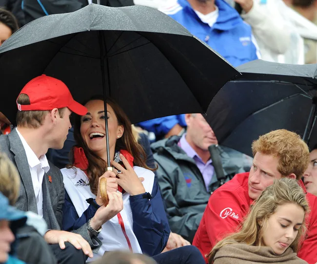 Group of people at an outdoor event under umbrellas, smiling and chatting, with one person wearing a red cap.