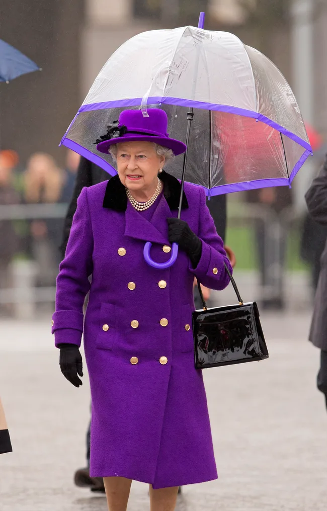 Elderly woman in a purple coat and hat holding a clear umbrella with a purple trim, walking outdoors.