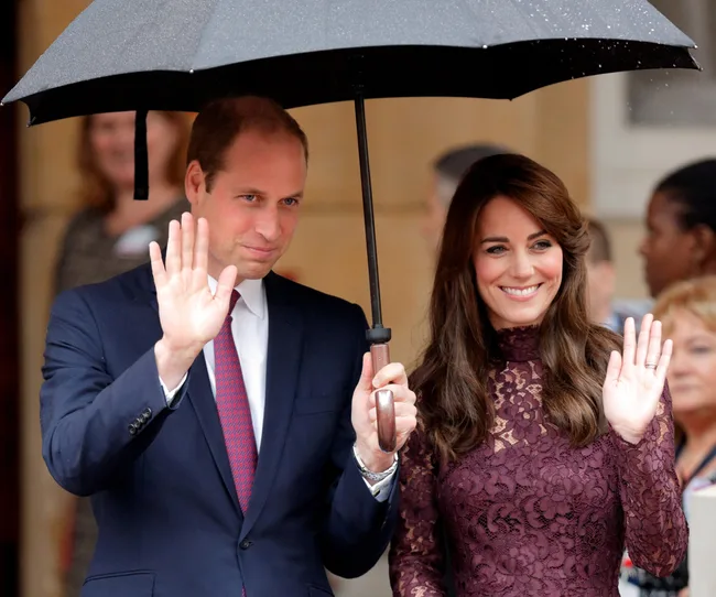 A man and woman smiling and waving under an umbrella, with people in the background.