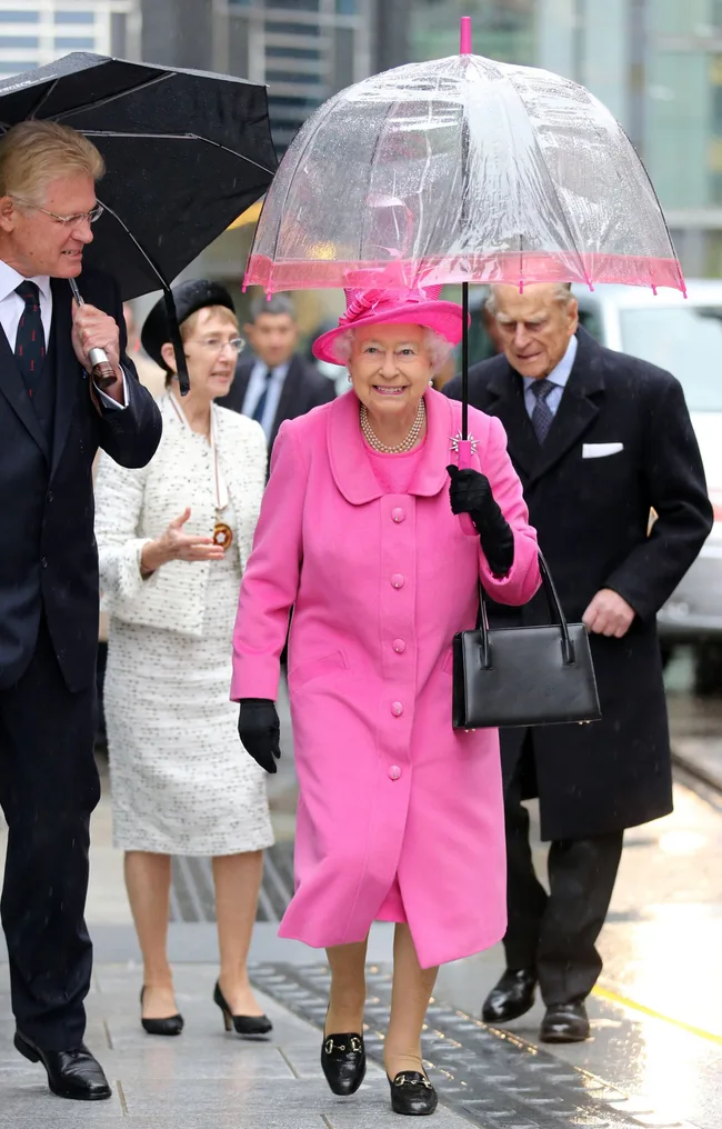 A woman in a bright pink coat and hat smiles while holding a clear umbrella, walking with companions in rainy weather.
