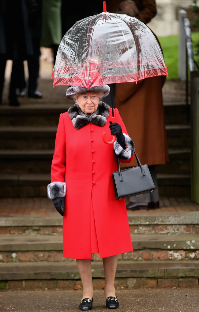 A person in a red coat and hat holds a transparent umbrella while standing outdoors.