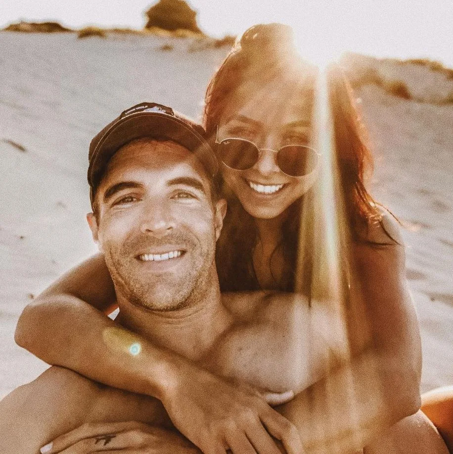 A couple smiling on a sunny beach, the man wearing a cap and the woman wearing sunglasses, embracing with sunlight behind them.