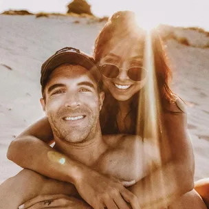 A couple smiling on a sunny beach, the man wearing a cap and the woman wearing sunglasses, embracing with sunlight behind them.