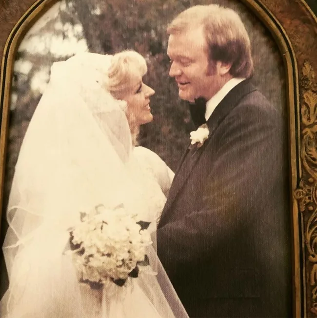 Bride and groom in a vintage wedding photo, smiling at each other with trees in the background.