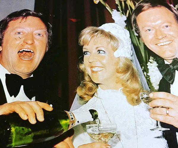 A bride and two men in tuxedos at a wedding, one pouring champagne, all smiling and looking joyful.