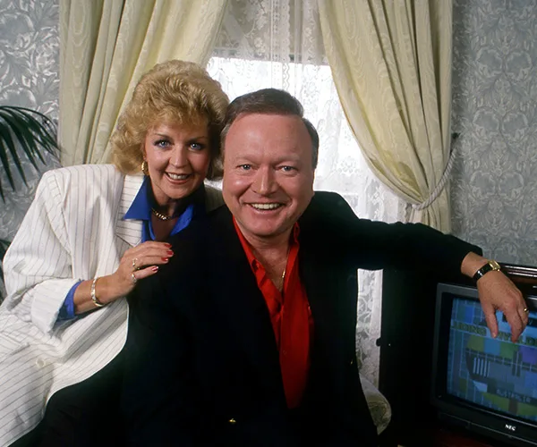 A man and woman smiling in a living room with a television in the background.