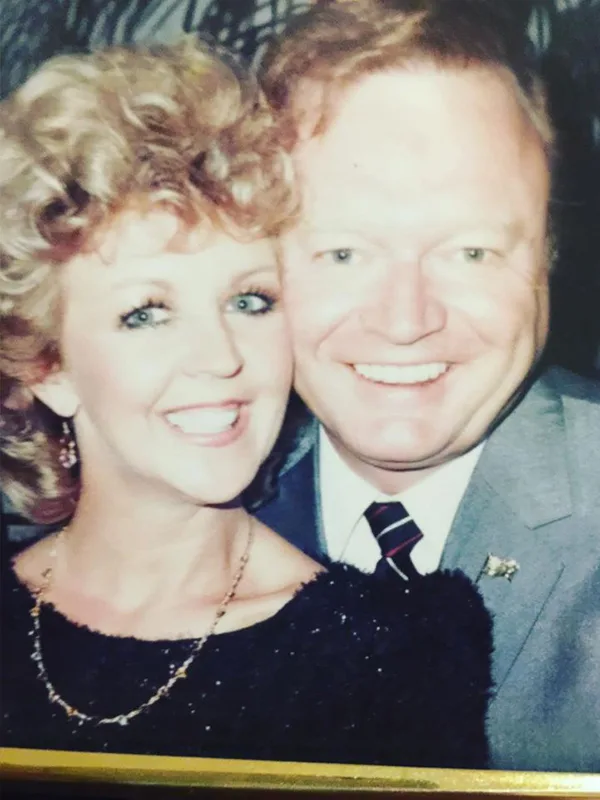 A smiling man and woman close together, indoors; man in suit and tie, woman in black top.