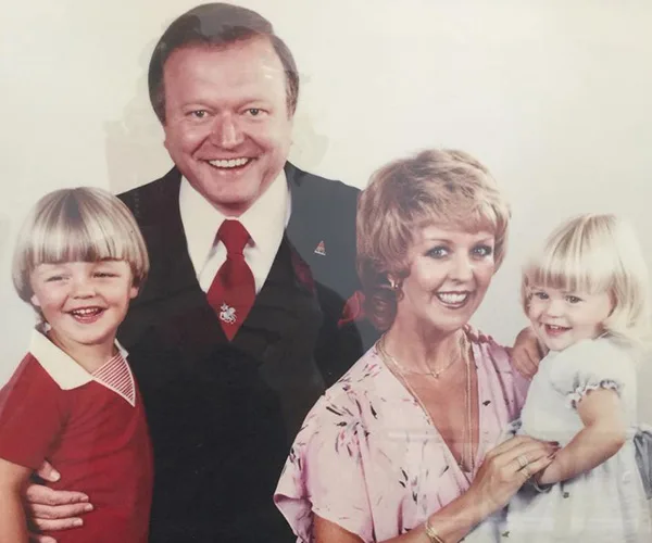 A smiling family portrait with two children and two adults, the man in a suit and tie, and the woman in a pink blouse.