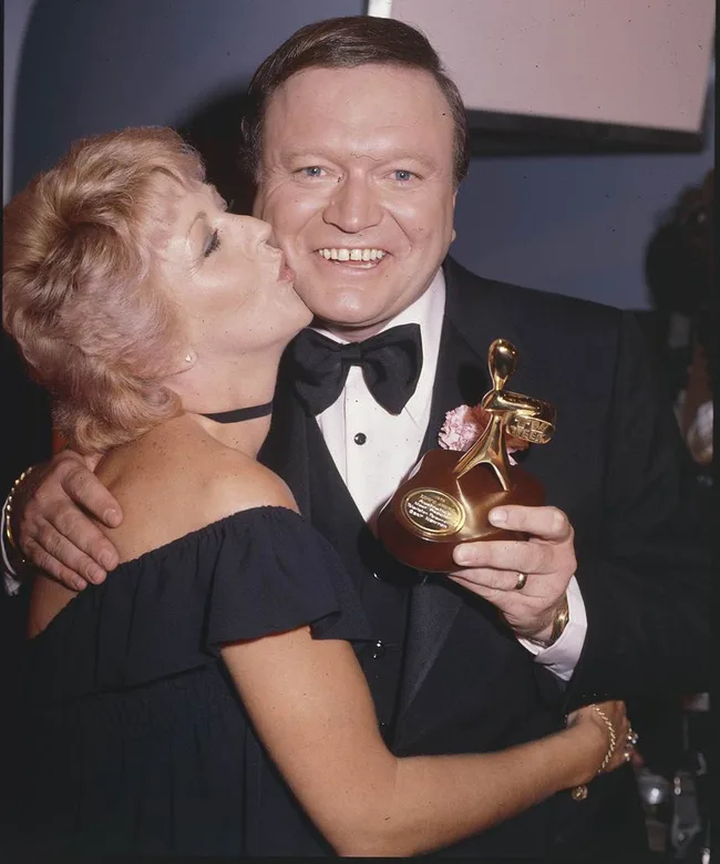 Concertgoer kisses man holding an award, both in formal wear, smiling at an event.