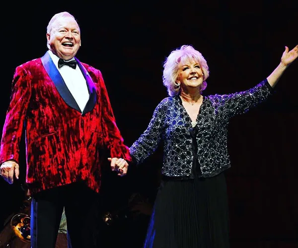 Elderly couple onstage, smiling and holding hands; the man in a red jacket, the woman in a sparkly black top.