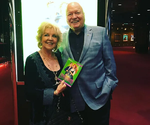 A woman and man smiling, holding a DVD, standing in a cinema lobby.