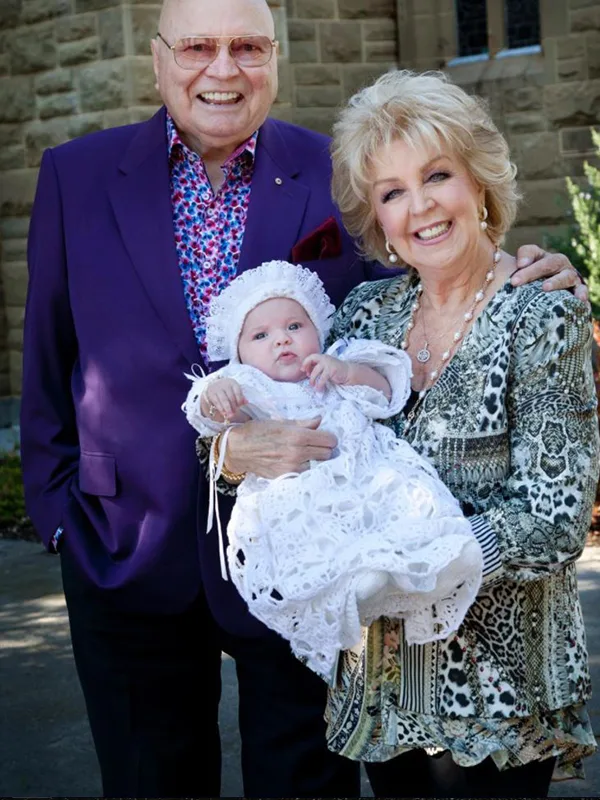An elderly couple smiling, holding a baby in a christening gown outside a stone building.