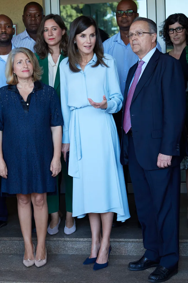 Group of people standing together, with a woman in a blue dress speaking, in an outdoor setting.