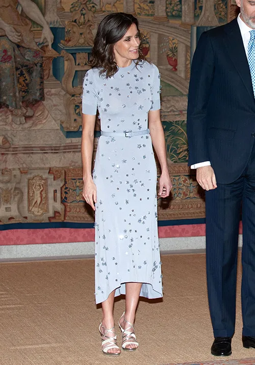 A woman in a light blue floral dress stands indoors beside a man in a dark suit, with an ornate tapestry background.