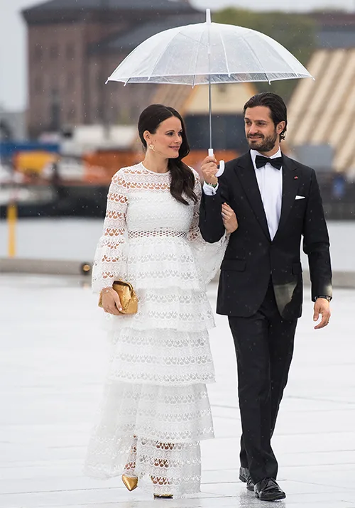 A couple elegantly dressed strolls under an umbrella; the woman in a white lace dress, the man in a black tuxedo.
