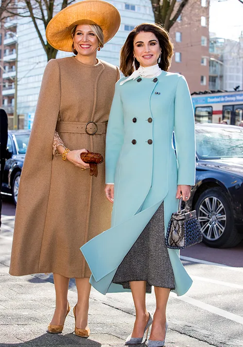 Two women in elegant coats, one in tan with a cape and wide-brim hat, the other in blue with a handbag, standing on a street.
