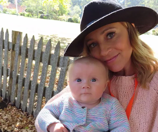 A woman in a hat holds a baby, both smiling, with a picket fence and garden in the background.