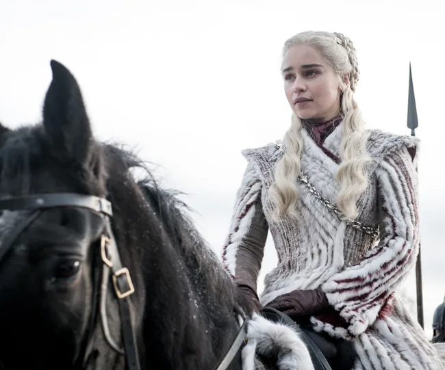 Daenerys Targaryen in a fur coat riding a black horse, with braided blonde hair, from Game of Thrones.