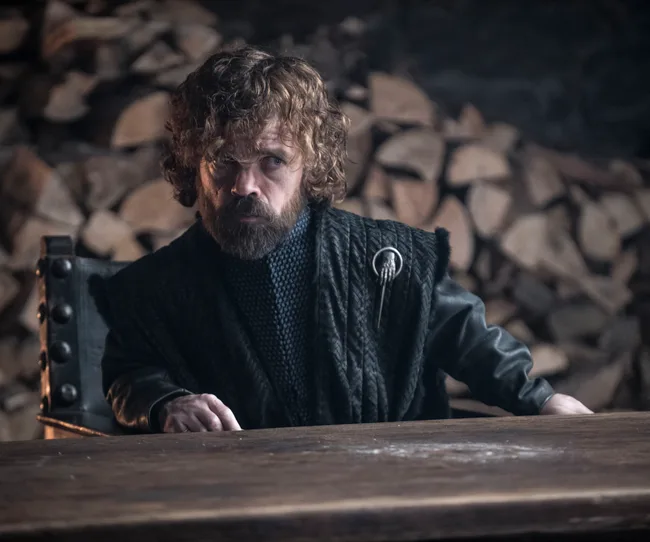 Tyrion Lannister from "Game of Thrones" sitting at a table, wearing dark clothing with wood logs in the background.