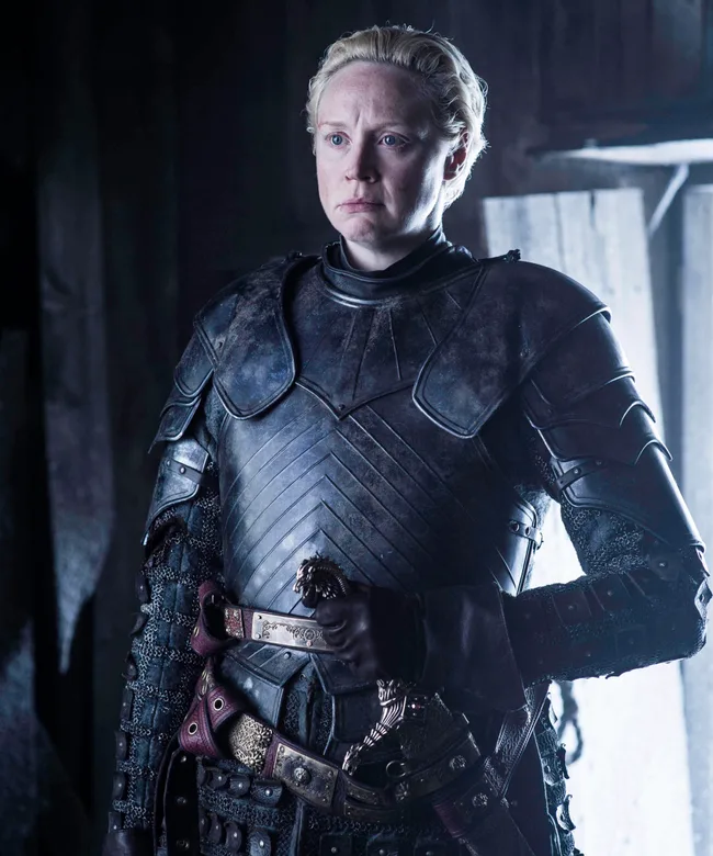 Brienne of Tarth in dark armor, stands inside a dimly lit room, appearing serious and focused.