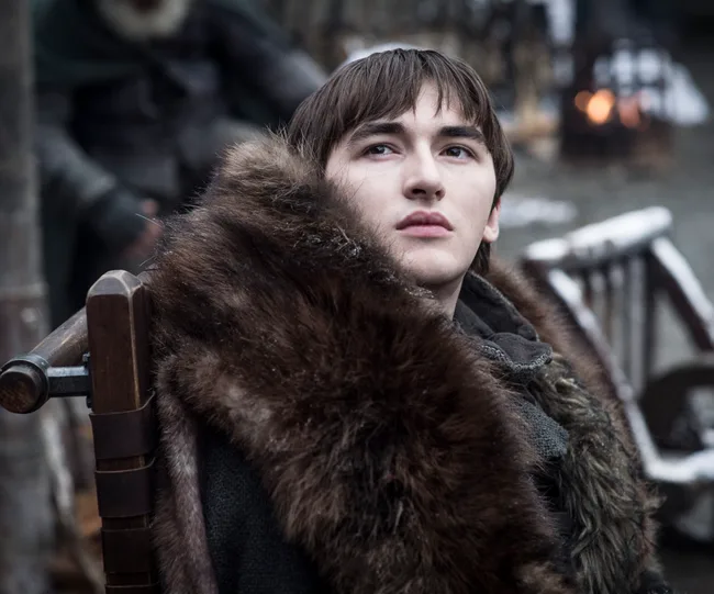 Bran Stark in fur coat sitting outdoors with a thoughtful expression, scene from "Game of Thrones."