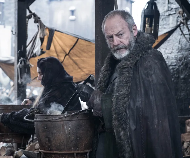 A bearded man in medieval attire stands by a pot, holding a ladle, outdoors. It's a scene from "Game of Thrones."