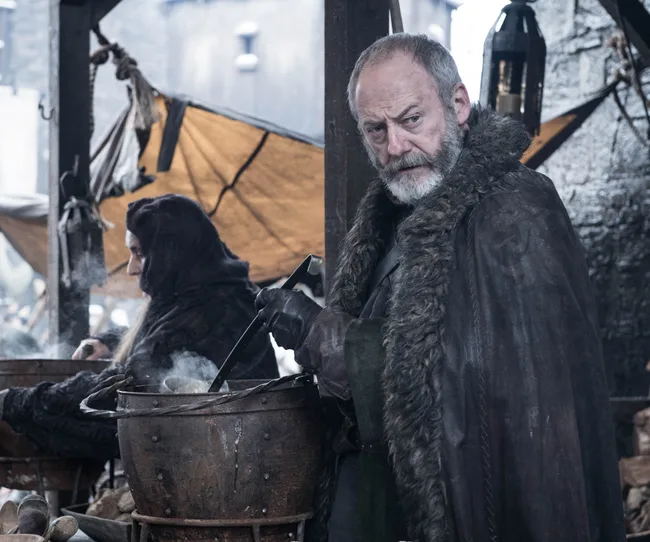A bearded man in medieval attire stands by a pot, holding a ladle, outdoors. It's a scene from "Game of Thrones."