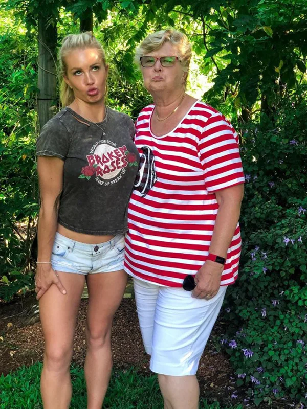 Two people making funny faces in a garden, one in a graphic tee and shorts, the other in a striped shirt and white shorts.