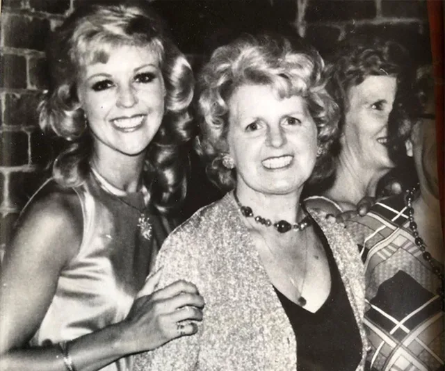 Three women smile in a black and white photo, wearing 1970s-style outfits and hairstyles.