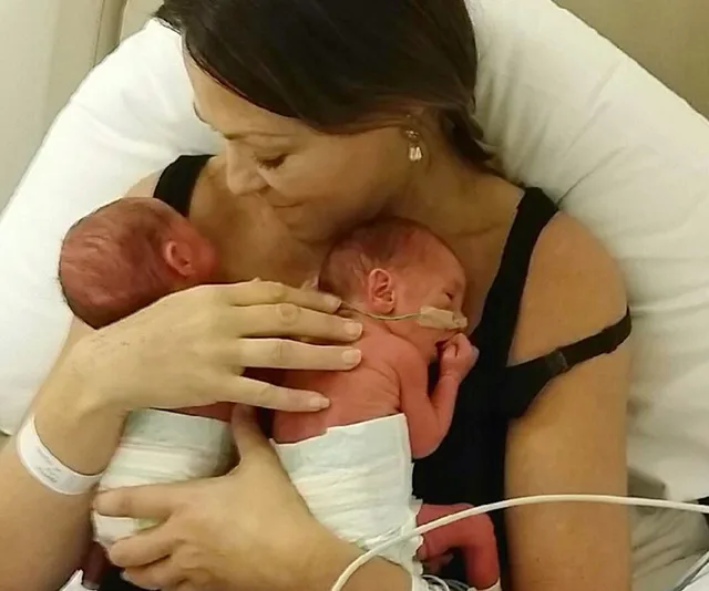 A woman lovingly holds two newborn twins wrapped in diapers, lying on her chest.