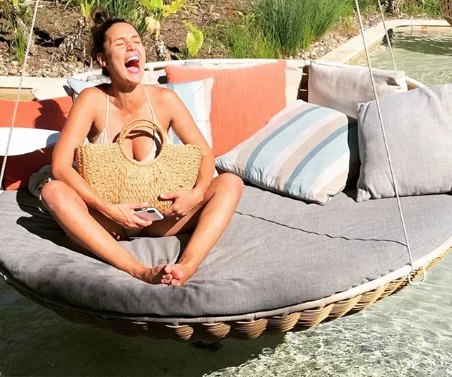 A woman in a bikini laughs while sitting on a circular swing over water, holding a straw bag and a phone.