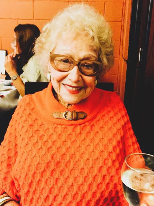Elderly woman with glasses, smiling, wearing an orange sweater, sitting at a restaurant table with a drink.