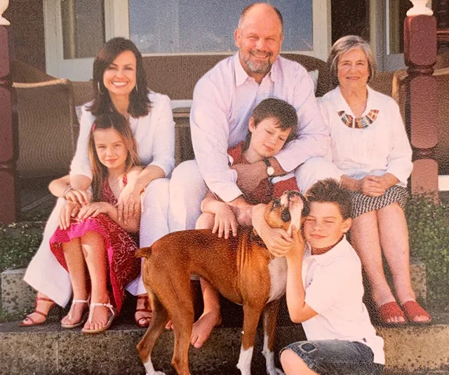 Family sitting on porch with three children, a man, two women, and a dog. Smiling faces and casual clothing.