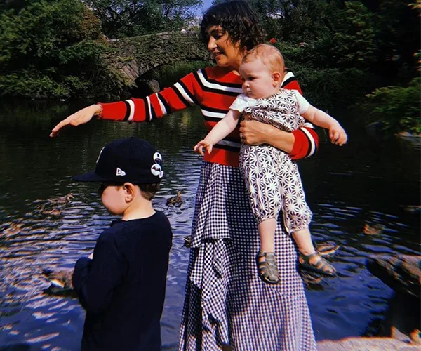 Woman in striped sweater holding a baby, pointing, with a child in a cap by a pond in a park setting.
