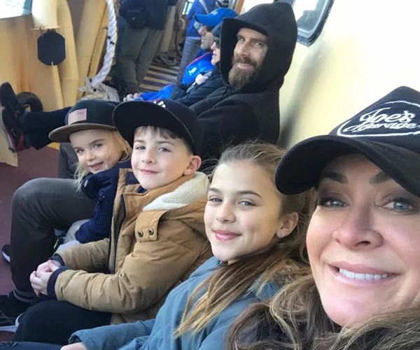 Family of four bundled up in jackets and hats, sitting closely on a boat, with a smiling woman taking a selfie.