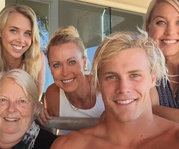 A group selfie with five smiling people, including a shirtless young man and four women, in a casual setting.
