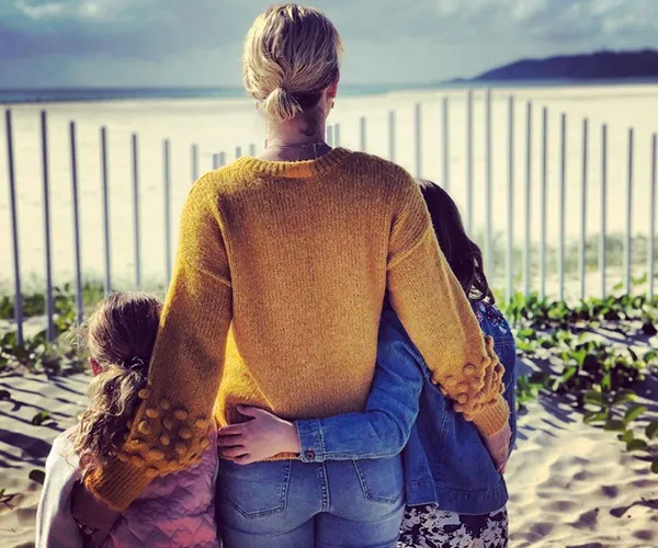 Woman in yellow sweater embraces two children at a sandy beach.