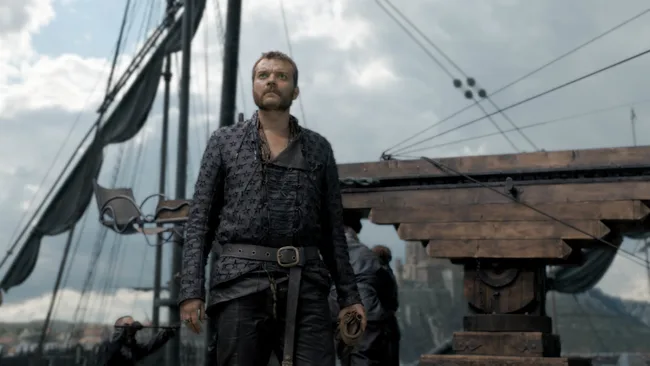 A man in medieval attire stands on a wooden ship deck, with sails and rigging visible in the background under a cloudy sky.