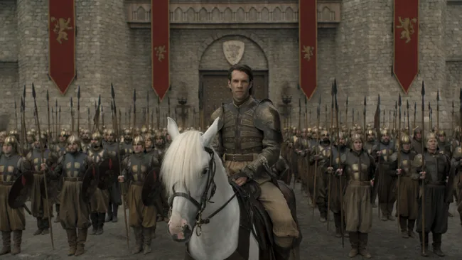 A man in armor rides a white horse in front of a formation of soldiers holding spears, standing outside a castle gate.